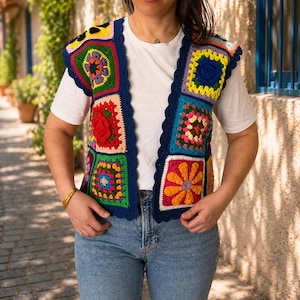 May include: A vibrant, patchwork crochet vest with a dark blue trim. The vest showcases diverse square patterns with floral and geometric designs in red, yellow, green, and blue. It is worn over a white t-shirt and blue jeans.