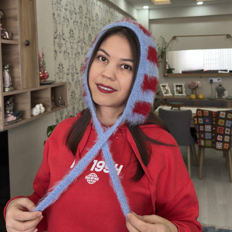 May include: A woman wearing a blue and red striped fuzzy knit hat with a long tie. The hat is tied in a knot at the front of her neck. She is wearing a red hoodie with the text "1994" printed on the front.