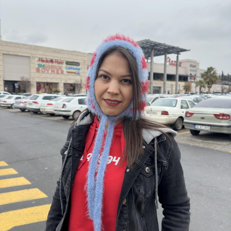 May include: A woman wearing a red and blue fuzzy knit hat with long ties. She is wearing a red hoodie with a white graphic and a black denim jacket.