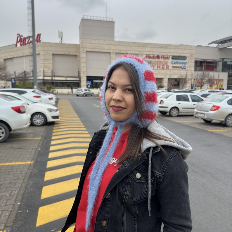 May include: A woman wearing a red and blue striped fuzzy hat with long blue strings. She is wearing a black denim jacket over a red shirt. The woman is standing in a parking lot in front of a shopping mall.