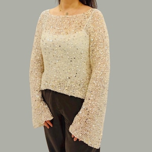 Può includere: Un maglione color crema a maniche lunghe con scollo a barchetta e maniche a campana. Il maglione è impreziosito da paillettes e ha un design a maglia larga e aperta. L'indumento è abbinato a pantaloni marrone scuro.
