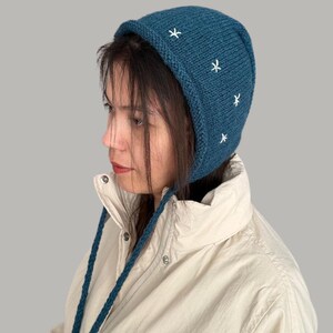 May include: A teal blue knitted hat with a long tie, featuring white embroidered stars. The hat has a pointed crown and a wide brim.