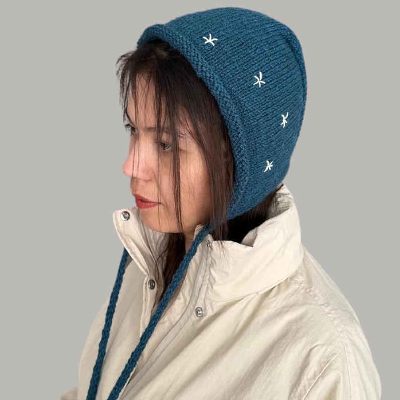 May include: A teal blue knitted hat with a long tie, featuring white embroidered stars. The hat has a pointed crown and a wide brim.