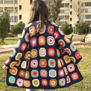 Peut inclure: Un long cardigan en crochet bleu marine avec un motif géométrique coloré de fleurs crochetées. Le cardigan est ample et a une longue ouverture sur le devant.