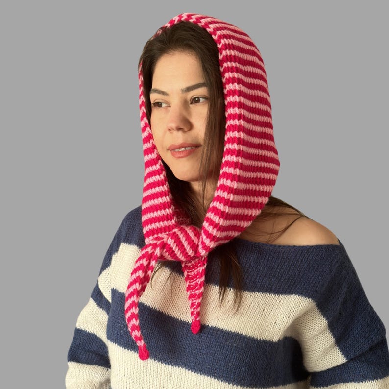 May include: A pink and white striped knitted hood with long ties, worn over a blue and white striped sweater. The hood is made of a soft, textured yarn and is designed to cover the head and neck.