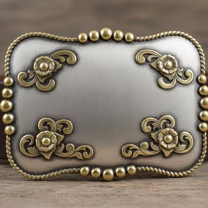 Op de afbeelding: Een rechthoekige zilverkleurige gesp met sierlijke goudkleurige bloemmotieven. De gesp heeft een touwachtige rand en goudkleurige balaccenten. Een bruine leren riem is zichtbaar achter de gesp.