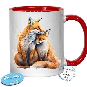 May include: White ceramic mug with a red rim and handle. The mug features a watercolour illustration of two red foxes cuddling. The text "Handmade by Just So Gifts" is printed on the mug.