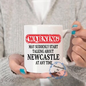 Peut inclure: Mug en c&eacute;ramique blanche avec le texte "WARNING MAY SUDDENLY START TALKING ABOUT NEWCASTLE AT ANY TIME" en noir et rouge. La tasse a une anse et est tenue par une personne. La tasse est faite &agrave; la main.