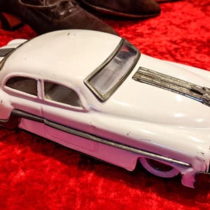 May include: A white vintage toy car with black and silver accents. The classic car is set against a red background. The toy car has a detailed design, including a front grill and side trim.