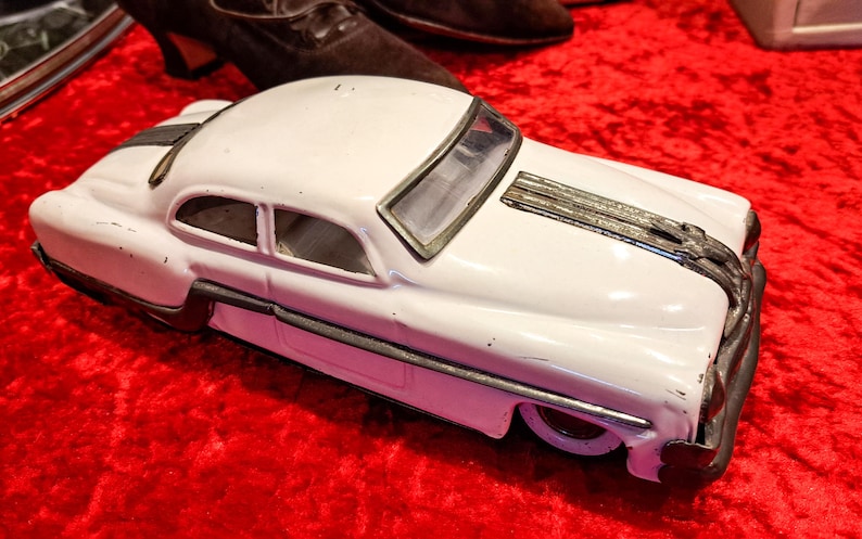 May include: A white vintage toy car with black and silver accents. The classic car is set against a red background. The toy car has a detailed design, including a front grill and side trim.