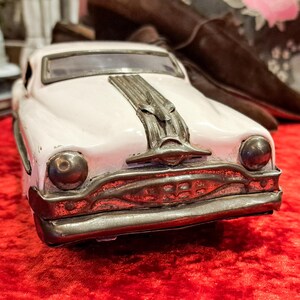 May include: A vintage-style toy car in a soft pink hue, featuring a detailed chrome grill and hood ornament. The car's design evokes classic automotive aesthetics. It sits on a vibrant red surface, with blurred objects in the background.