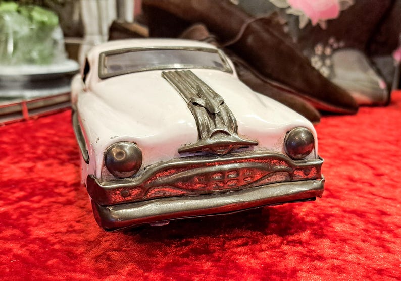 May include: A vintage-style toy car in a soft pink hue, featuring a detailed chrome grill and hood ornament. The car's design evokes classic automotive aesthetics. It sits on a vibrant red surface, with blurred objects in the background.