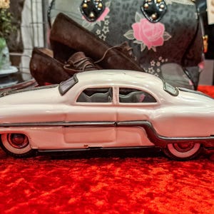 May include: A white vintage toy car with black trim and red wheels sits on a red surface. The classic car has a streamlined design and is a collectible item. The car is a coupe style.
