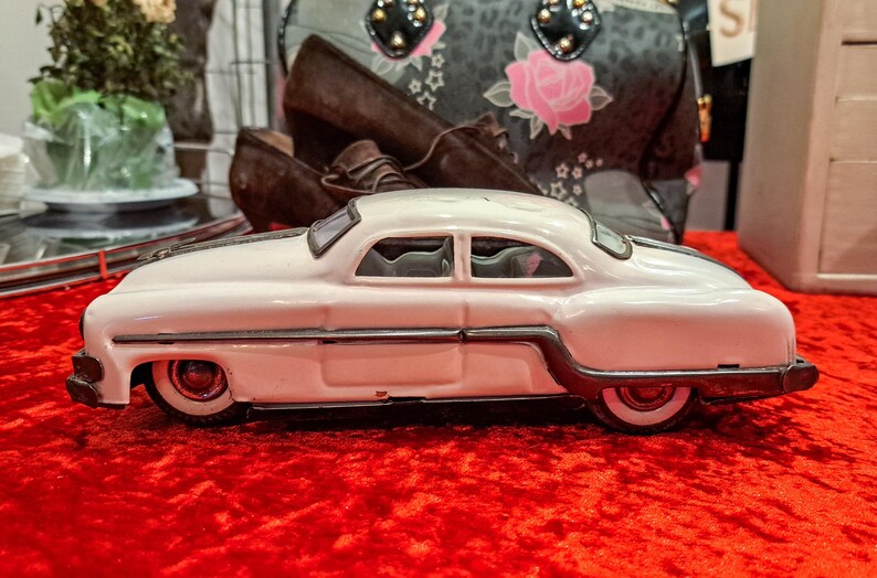May include: A white vintage toy car with black trim and red wheels sits on a red surface. The classic car has a streamlined design and is a collectible item. The car is a coupe style.