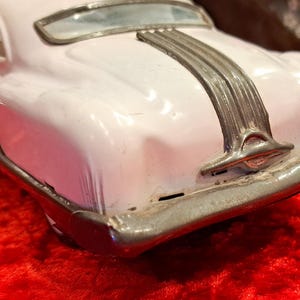 May include: A vintage toy car in a light pink color, with a silver-colored trim and accents. The car's rear features a detailed bumper and a decorative silver stripe. The toy car is a classic collectible item.