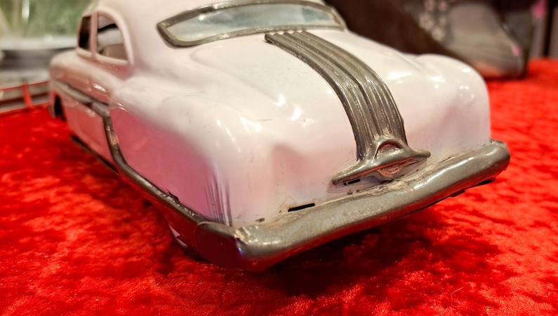 May include: A vintage toy car in a light pink color, with a silver-colored trim and accents. The car's rear features a detailed bumper and a decorative silver stripe. The toy car is a classic collectible item.