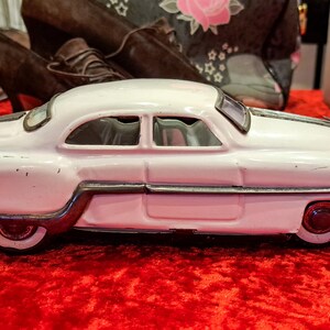 May include: A vintage white toy car with a silver trim and red wheels sits on a red surface. The classic car has a streamlined design with two windows and a black stripe on the roof. The toy car is a collectible item.