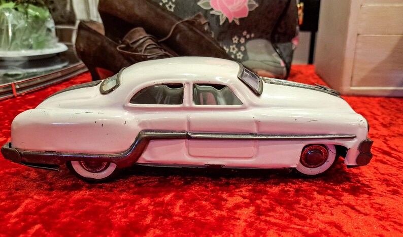 May include: A vintage white toy car with a silver trim and red wheels sits on a red surface. The classic car has a streamlined design with two windows and a black stripe on the roof. The toy car is a collectible item.