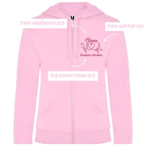 Peut inclure: Un sweat à capuche rose clair avec fermeture éclair et poches avant. Le sweat arbore un motif bordeaux avec un tracé de battement de cœur, un stéthoscope et le nom "Clara". Le texte "Étudiante infirmière" est également présent.
