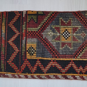 May include: Rectangular woven rug with a geometric pattern in black, red, yellow, and green. The rug has a fringed edge and a repeating diamond and zigzag design. The rug is likely handmade.