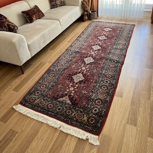 May include: A red, black, and cream patterned rug with fringe on the edges. The rug is laid out on a hardwood floor in a living room setting.