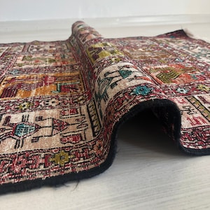 May include: A rolled up rug with a colorful geometric pattern in shades of red, orange, green, blue, and black. The rug has a black fringe border.