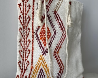 Può includere: Una borsa tote bianca con un designo tessuto in tonalità di rosso, arancione, giallo e viola. La borsa ha due manici in corda bianca con nappe.