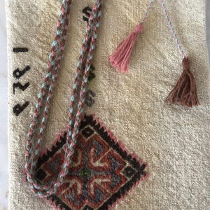 May include: A beige fabric bag with a brown and blue braided strap and pink and brown tassels. The bag has a diamond-shaped embroidered design with red, green, and black accents. The year "1882" is embroidered on the bag.