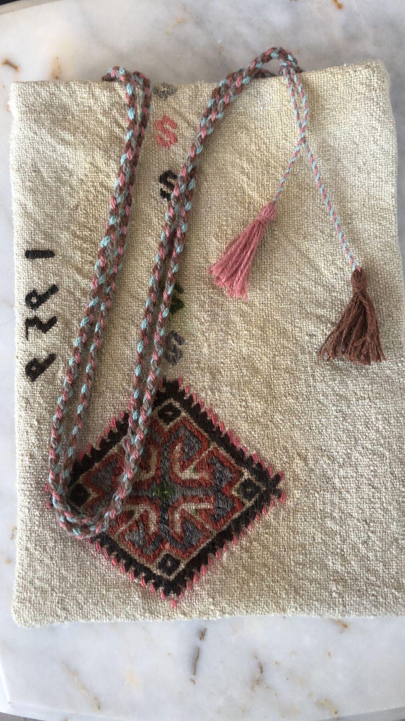May include: A beige fabric bag with a brown and blue braided strap and pink and brown tassels. The bag has a diamond-shaped embroidered design with red, green, and black accents. The year "1882" is embroidered on the bag.