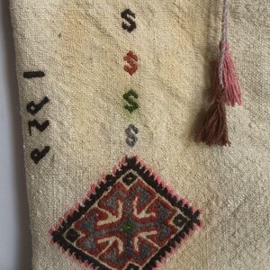 May include: A white fabric pouch with a brown, pink, and blue tassel. The pouch has a diamond-shaped embroidered design in shades of red, blue, green, and black. The pouch has the number "1221" written on it.
