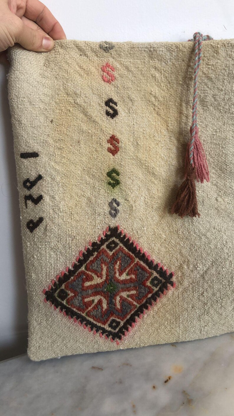 May include: A white fabric pouch with a brown, pink, and blue tassel. The pouch has a diamond-shaped embroidered design in shades of red, blue, green, and black. The pouch has the number "1221" written on it.