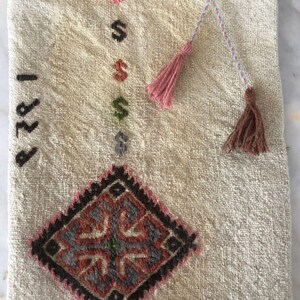 May include: A white woven tote bag with a brown and red diamond pattern and the number "1989" embroidered on the front. The bag has two pink and brown tassels attached to the handles.