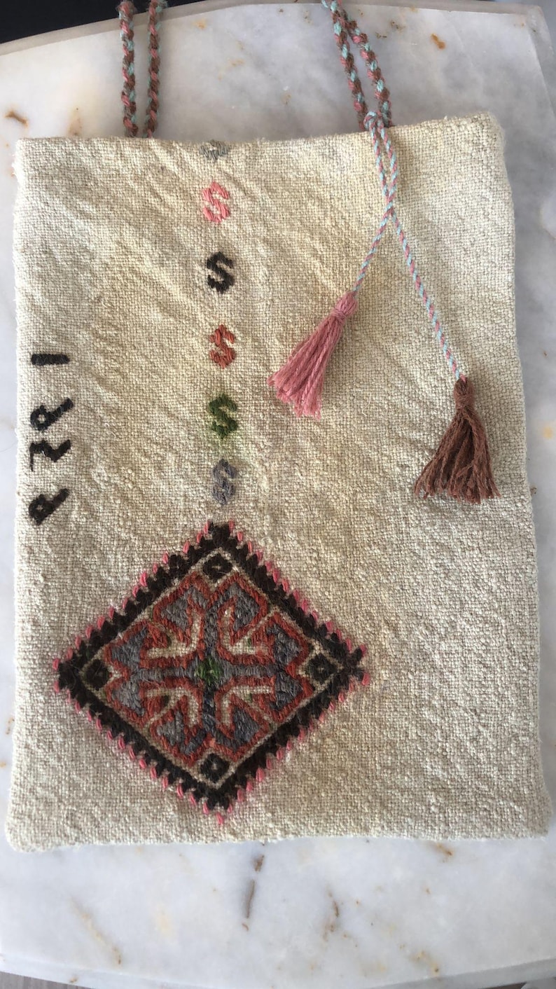 May include: A white woven tote bag with a brown and red diamond pattern and the number "1989" embroidered on the front. The bag has two pink and brown tassels attached to the handles.