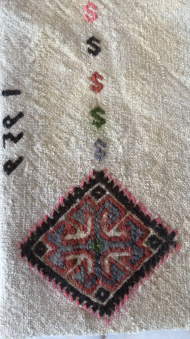 May include: A white fabric with a black and red embroidered design. The design is a diamond shape with a floral pattern in the center. The fabric has the number "1978" written on it in black thread.