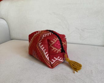 May include: A small red and pink patterned zippered pouch with a gold tassel. The pouch is made of woven fabric and has a geometric design.