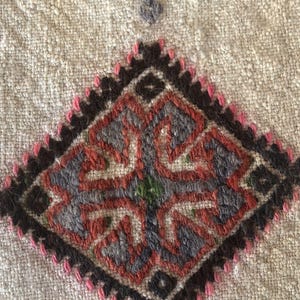 May include: Close-up of a beige fabric with a diamond-shaped embroidered design. The design features a central motif with four arms, each with a smaller diamond shape. The embroidery is done in shades of red, black, gray, and green.