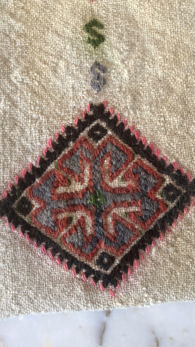 May include: Close-up of a beige fabric with a diamond-shaped embroidered design. The design features a central motif with four arms, each with a smaller diamond shape. The embroidery is done in shades of red, black, gray, and green.
