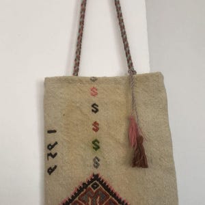 May include: A beige woven tote bag with a brown and pink tassel. The bag has a geometric design embroidered in red, brown, green, and blue. The number "1828" is embroidered on the side of the bag.