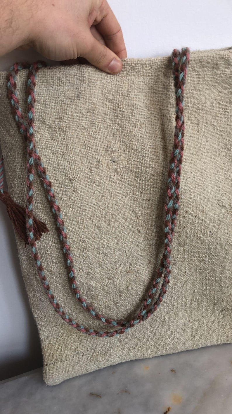 May include: A beige tote bag with two braided straps. The straps are made of brown, pink, and blue yarn.