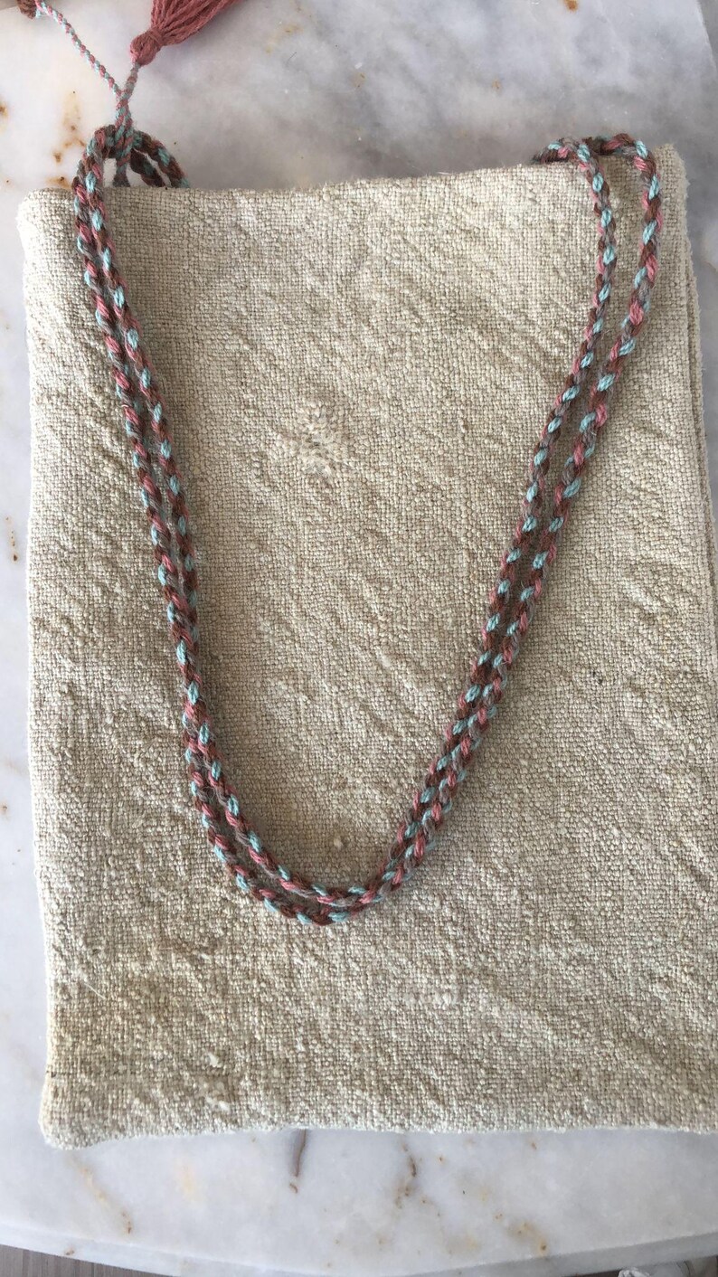May include: A beige linen tote bag with a braided strap made of brown, pink, and blue yarn.