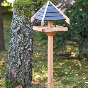 Può includere: Una mangiatoia per uccelli in legno con un tetto a punta grigio scuro e un vassoio di alimentazione quadrato. La mangiatoia è montata su un lungo palo quadrato di legno. Lo sfondo include un tronco d'albero e fogliame verde.