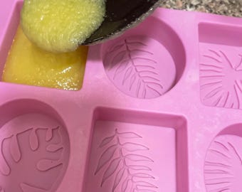 May include: A pink silicone mold with leaf designs is being filled with yellow liquid soap from a black ladle. The mold has square and round cavities. Soap making process.