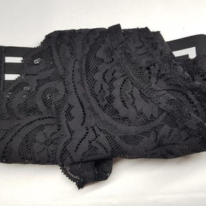 May include: Black lace fabric with an intricate floral pattern. The fabric is folded and appears to be part of a garment or trim. The lace has a delicate, open weave design, and the overall look is elegant and detailed.