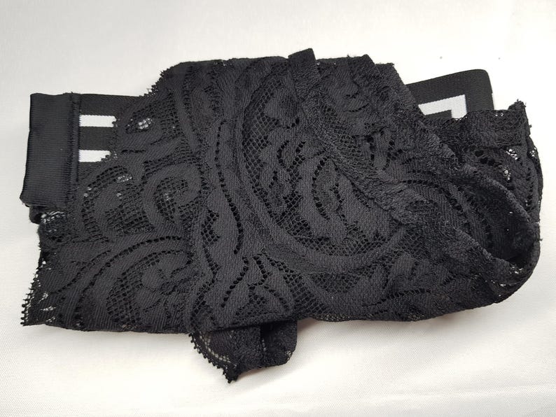 May include: Black lace fabric with an intricate floral pattern. The fabric is folded and appears to be part of a garment or trim. The lace has a delicate, open weave design, and the overall look is elegant and detailed.