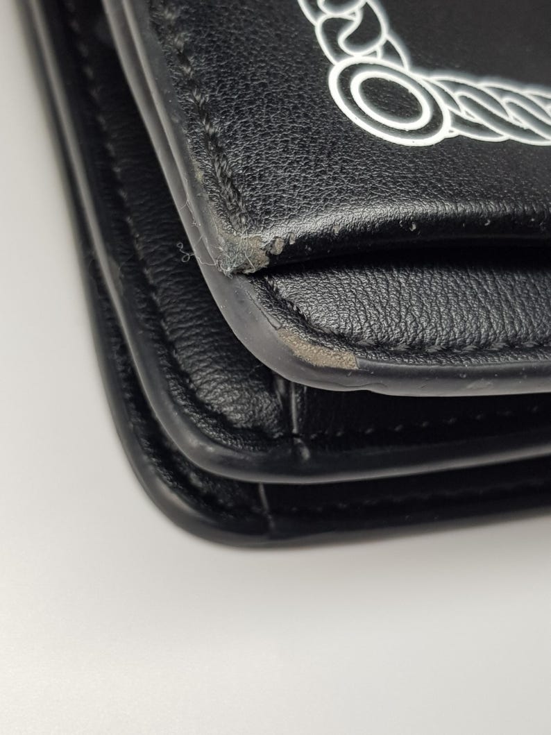 Puede incluir: Primer plano de un bolso de mano de cuero negro con un dise&ntilde;o de cadena blanca en la solapa. El bolso muestra signos de desgaste en los bordes. El bolso es probablemente un bolso de hombro o bandolera.