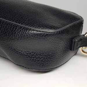 Pu&ograve; includere: Primo piano di una borsa in pelle nera con superficie strutturata. La borsa ha una forma arrotondata e una chiusura color oro attaccata a un cinturino in pelle nera. La borsa &egrave; su una superficie bianca.
