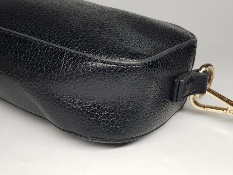 Pu&ograve; includere: Primo piano di una borsa in pelle nera con superficie strutturata. La borsa ha una forma arrotondata e una chiusura color oro attaccata a un cinturino in pelle nera. La borsa &egrave; su una superficie bianca.