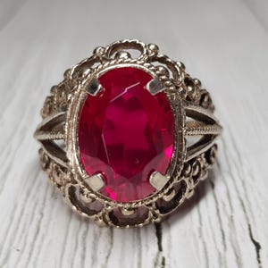 May include: Silver-toned ring with a large, oval-shaped, ruby-red gemstone. The ring features an ornate, filigree design around the gemstone and on the band. The ring is set against a white, textured surface.