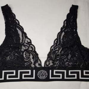 May include: Black lace bralette with a V-neck design. The bralette features a scalloped edge and a wide band with a white Greek key pattern and a circular emblem. The intricate lace detailing adds a touch of elegance to this lingerie piece.
