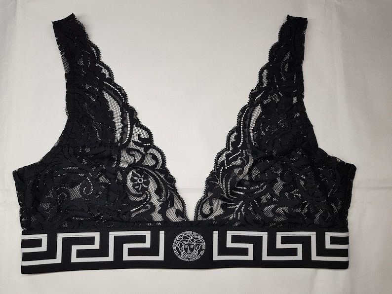 May include: Black lace bralette with a V-neck design. The bralette features a scalloped edge and a wide band with a white Greek key pattern and a circular emblem. The intricate lace detailing adds a touch of elegance to this lingerie piece.